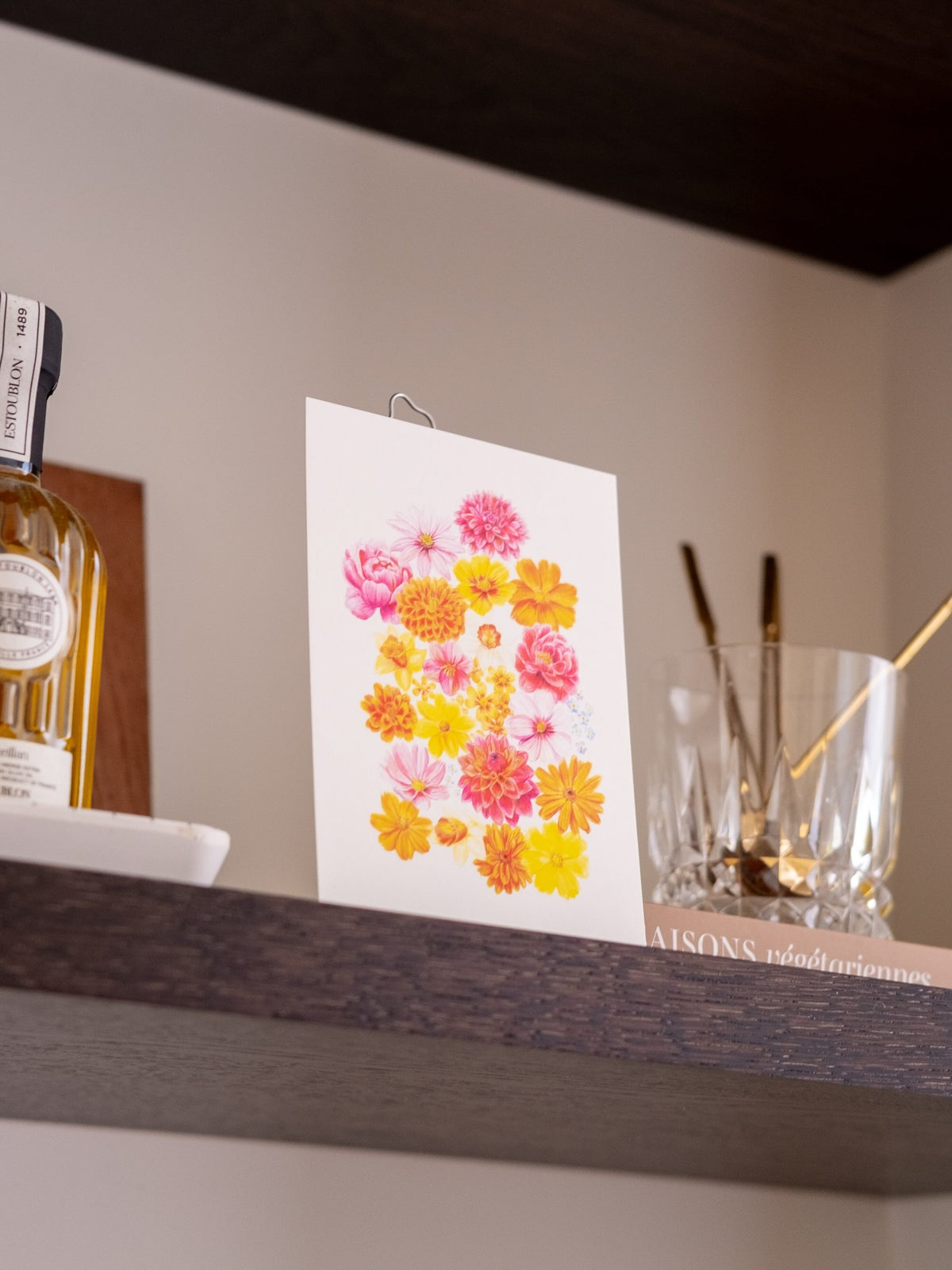 Carte postale « La Belle Saison » aux fleurs multicolores, présentée sur une étagère près d’un verre et de couverts.