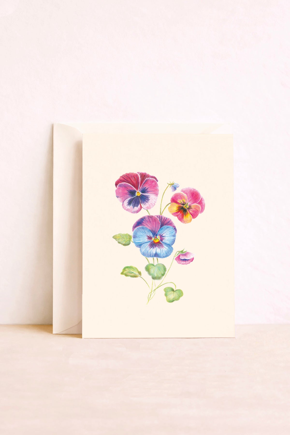 Carte postale illustrée de pensées roses, violettes, bleues et jaunes sur fond crème, avec une enveloppe derrière.