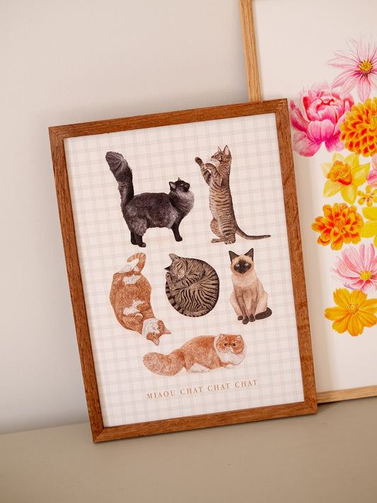 Affiche illustrée de six chats sur fond à carreaux, présentée dans un cadre en bois près de fleurs.