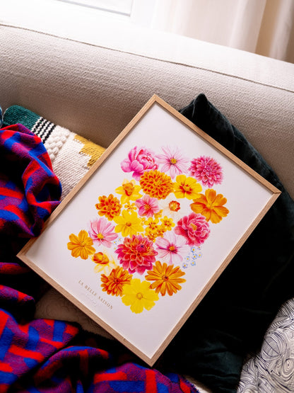 ffiche de fleurs « La Belle Saison », encadrée et posée sur un canapé parmi des coussins et un plaid coloré.
