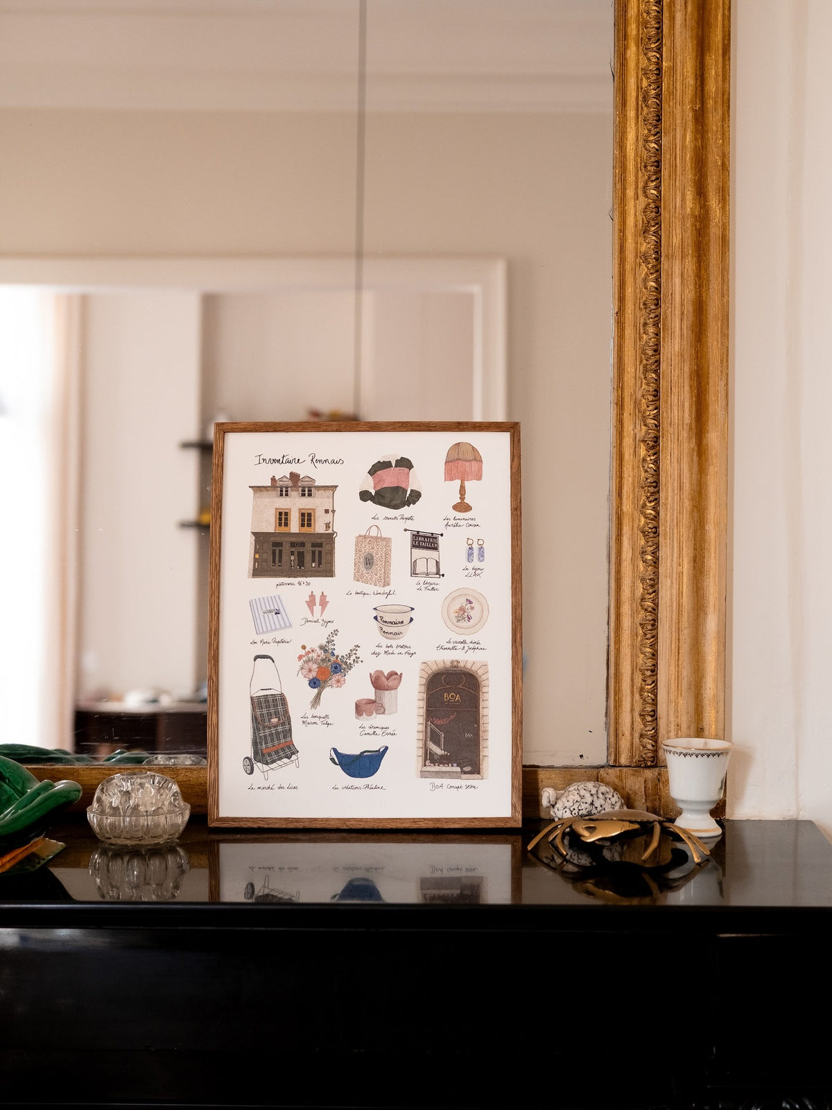 Affiche illustrée Inventaire Rennais encadrée, représentant une sélection de boutiques et d’objets rennais, posée sur une commode noire devant un miroir ancien.