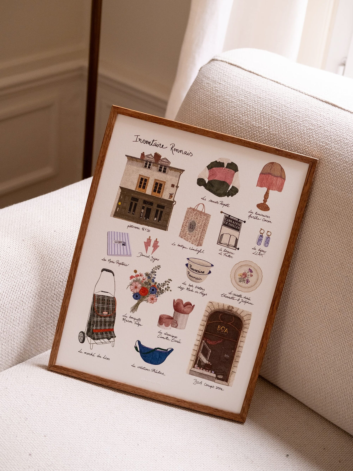 Affiche Inventaire rennais dans un cadre en bois, posée sur un fauteuil écru, avec ses illustrations de Rennes.