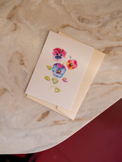 Carte postale Pensées aux fleurs multicolores et feuilles vertes, posée sur une enveloppe crème sur du marbre clair.