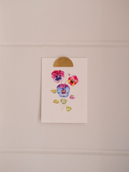 Carte postale illustrée de pensées multicolores, fixée sur un mur clair avec une pince dorée.