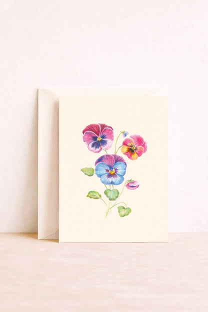 Carte postale illustrée de pensées roses, violettes, bleues et jaunes sur fond crème, avec une enveloppe derrière.