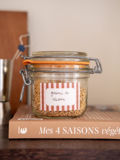 Étiquette adhésive à rayures rouges et blanches, marquée « graines de sésame », collée sur un bocal en verre.